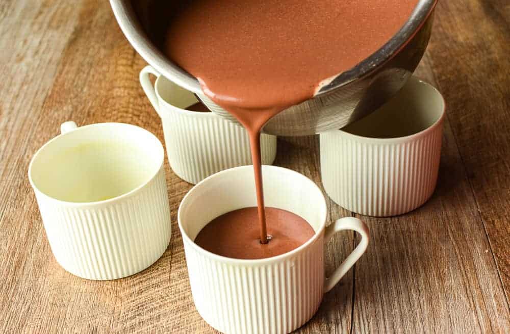 four white teacups on wooden table and chocolate being poured into one of the teacups from a stainless steel saucepan