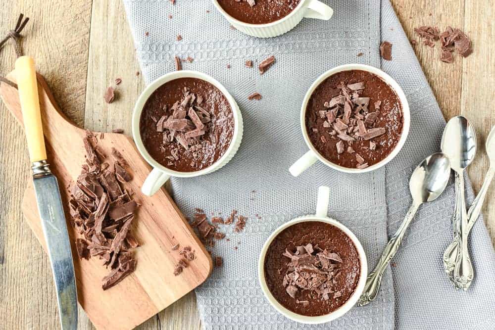 four teacups filled with chocolate pudding on a grey cloth with a wooden board of chopped chocolate viewed from above