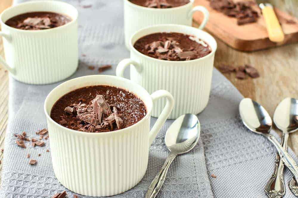 four teacups filled with chocolate pudding on a grey cloth with a wooden board of chopped chocolate and four silver spoons viewed from front on