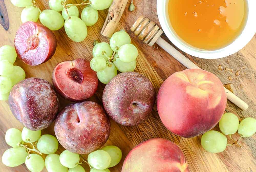 plums, peaches, green grapes, bowl of honey, honey twizzle stick, cinnamon stick and cardamom pods on timber board viewed from above