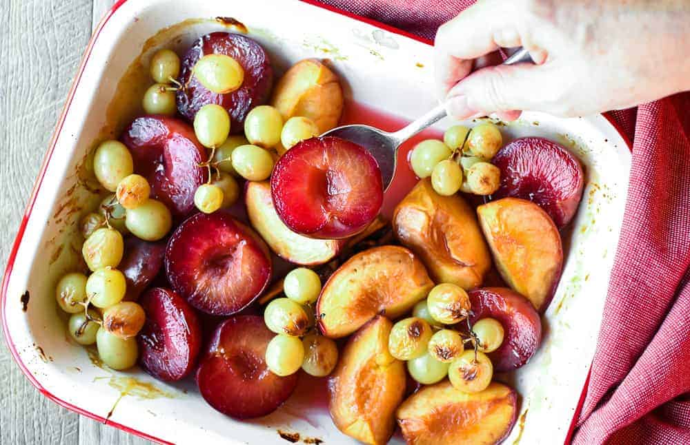Roasted Fruit with Prosecco and Honey is an easy dessert with vanilla ice cream and leftovers are particularly good for breakfast the next day. #fruit #summerfruit #stonefruit #grapes #roastedfruit #roasted #dessert #breakfast roasted plums, peaches and green grapes in white pan with red trim also hand spooning out plums viewed from above
