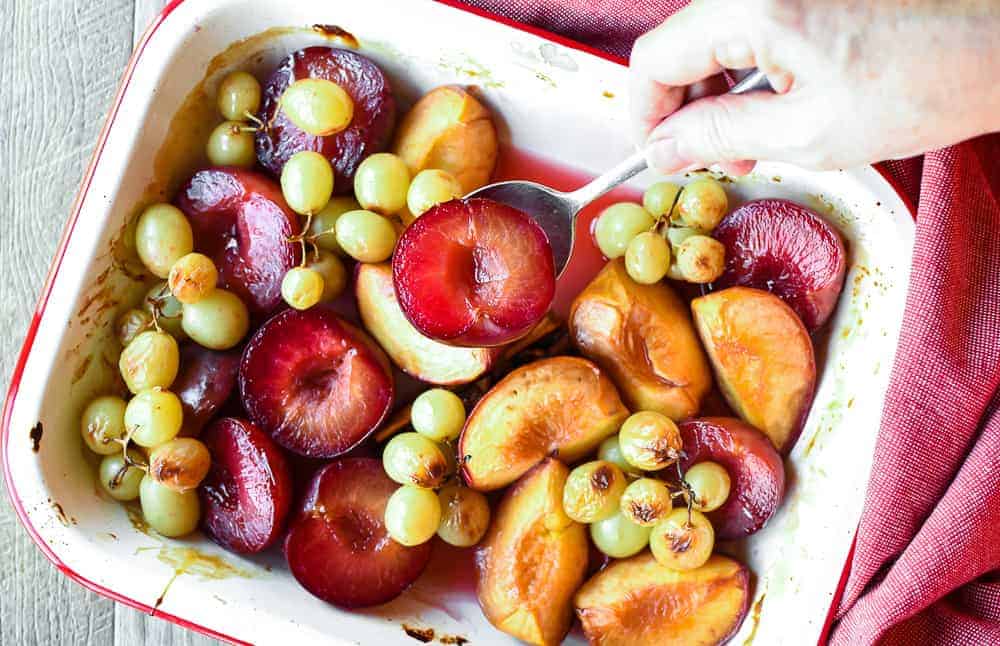 roasted plums, peaches and green grapes in white pan with red trim also hand spooning out plums viewed from above