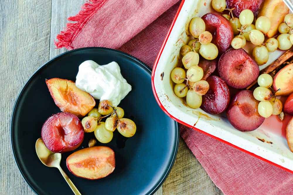 roasted plums, peaches and green grapes with scoop of yogurt on dark plate with gold spoon, on the right is a white pan with red trim filled with roasted plums, peaches and green grapes on red cloth. All is on a timber table and viewed from above