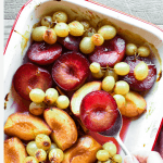 roasted plums, peaches and green grapes in white pan with red trim also hand spooning out plums viewed from above
