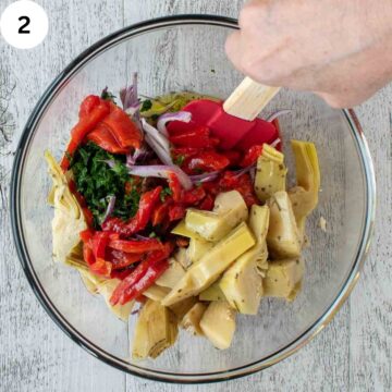 Artichoke hearts and other salad ingredients being mixed in a glass bowl.