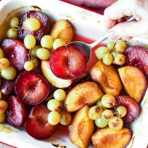 roasted plums, peaches and green grapes in white pan with red trim also hand spooning out plums viewed from above