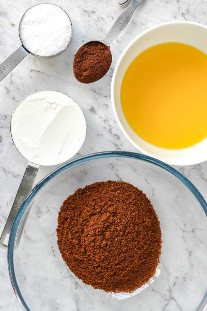 bowl of chocolate cookie crumbs, white bowl of melted butter, measuring cup filled with flour, measuring cup filled with sugar and spoon of cocoa