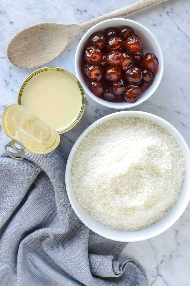 white bowl of coconut, white bowl of candied cherries, opened can of condensed milk, wooden spoon and grey cloth viewed from above