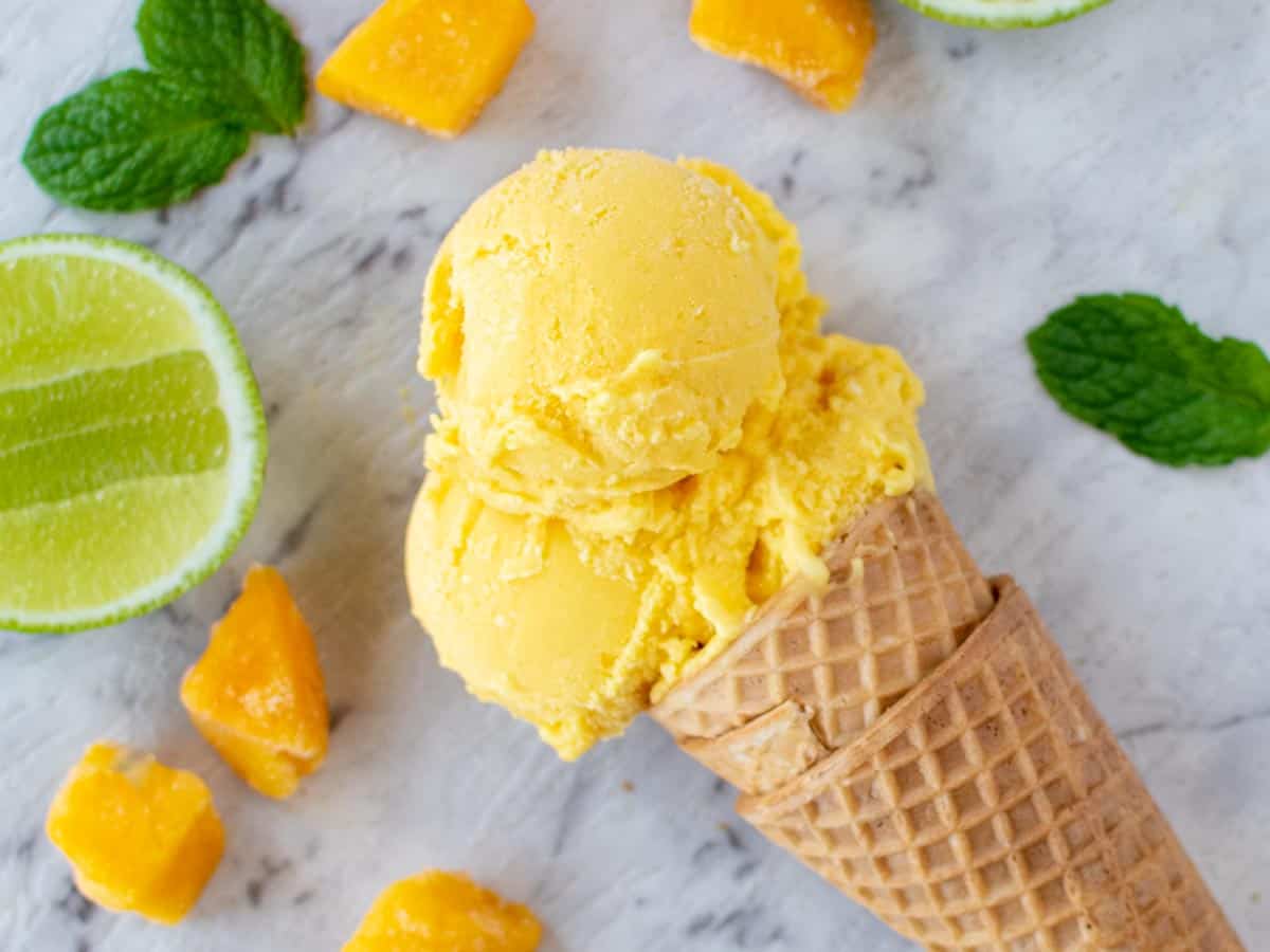 Close up of ice cream cone with 3 scoops on a marble table, chunks of mango, lime cheek and mint leaves.