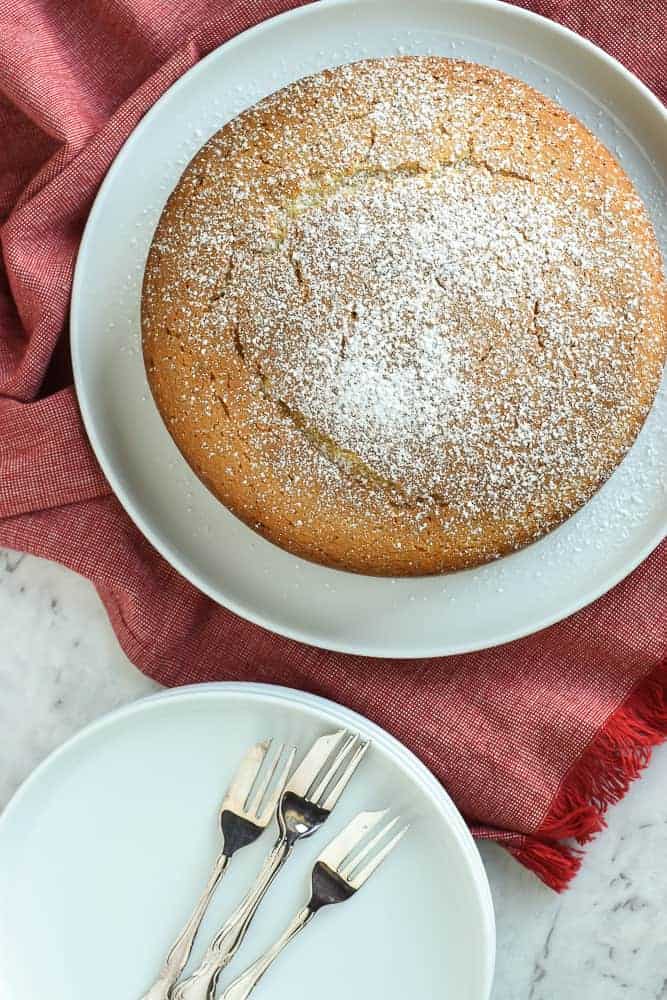 round vanilla cake dusted with powdered sugar on white plate on red cloth with white small plate and forks on the side viewed from above.