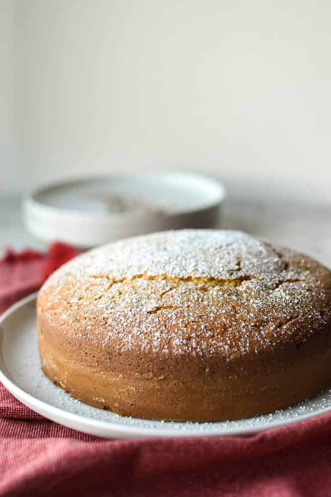 round plain cake dusted with powdered sugar on a white plate on a red cloth with white plates and forks in the background.