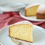 slice of plain cake on white plate with fork on red cloth with whole cake and extra plates in the background