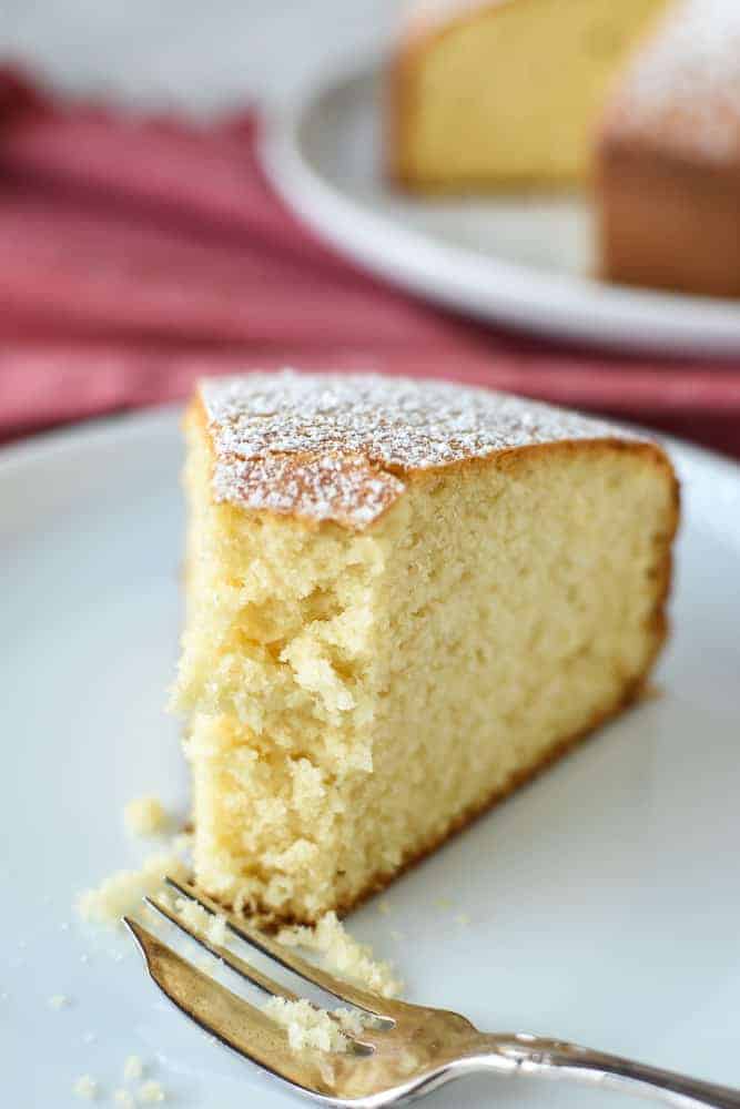 half eaten slice of plain cake on white plate with fork on red cloth with whole cake in the background.