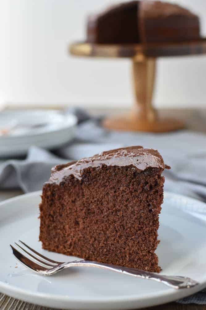 Slice of chocolate cake on white plate with cake fork, whole cake in the background.