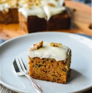 Square of carrot cake with cream cheese frosting on white plate with silver cake fork, remaining cake in background