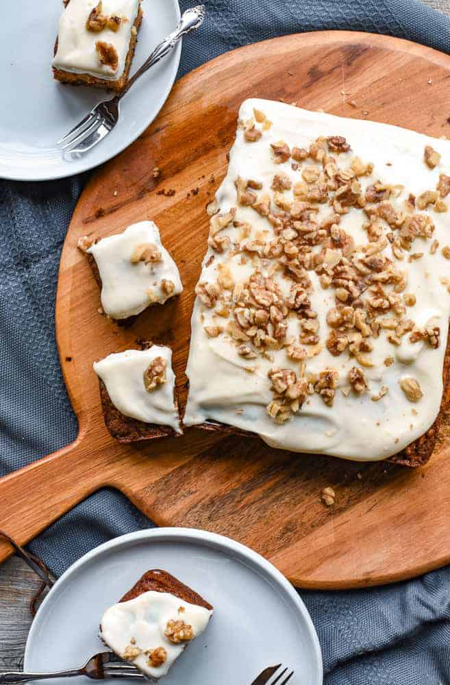 Carrot cake with cream cheese frosting and chopped walnuts on round wooden board with two squares of cake beside. Two white plates on either side with square of cake on.