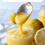 Lemon curd in glass jar with spoon pouring in lemon curd. In the backgroup cut and whole lemons