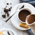 three cups and saucers of hot chocolate and hot chocolate being poured out of a white jug