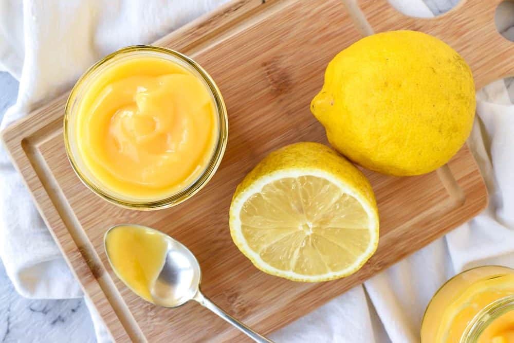 Homemade lemon curd is a tangy lemon custard which is perfect for cake fillings, tarts or to top scones. #homemade #lemoncurd #lemonrecipe #curdrecipe #homemadecurd Lemon curd in a jar, spoon, half a lemon and a whole lemon on a wooden board viewed from above.