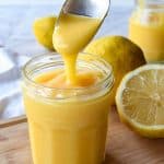 Lemon curd in glass jar with spoon pouring in lemon curd. In the backgroup cut and whole lemons