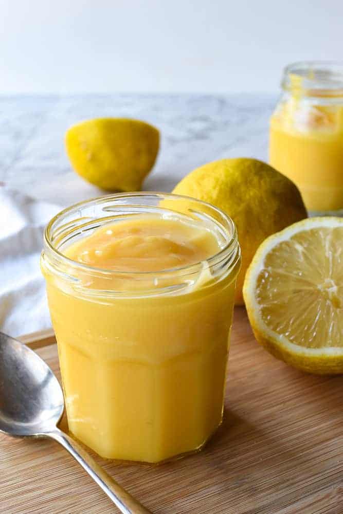 Homemade lemon curd is a tangy lemon custard which is perfect for cake fillings, tarts or to top scones. #homemade #lemoncurd #lemonrecipe #curdrecipe #homemadecurd Lemon curd in glass jar with spoon on timber board. In the backgroup cut and whole lemons