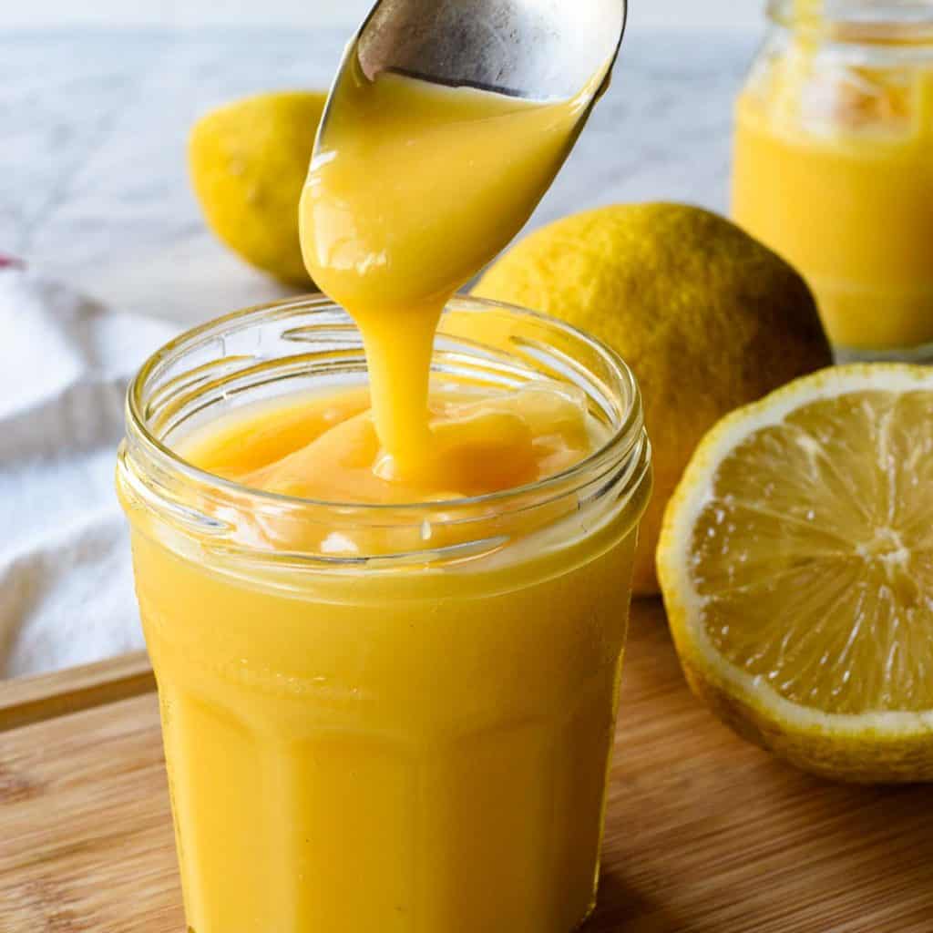Lemon curd in glass jar with spoon pouring in lemon curd. In the backgroup cut and whole lemons