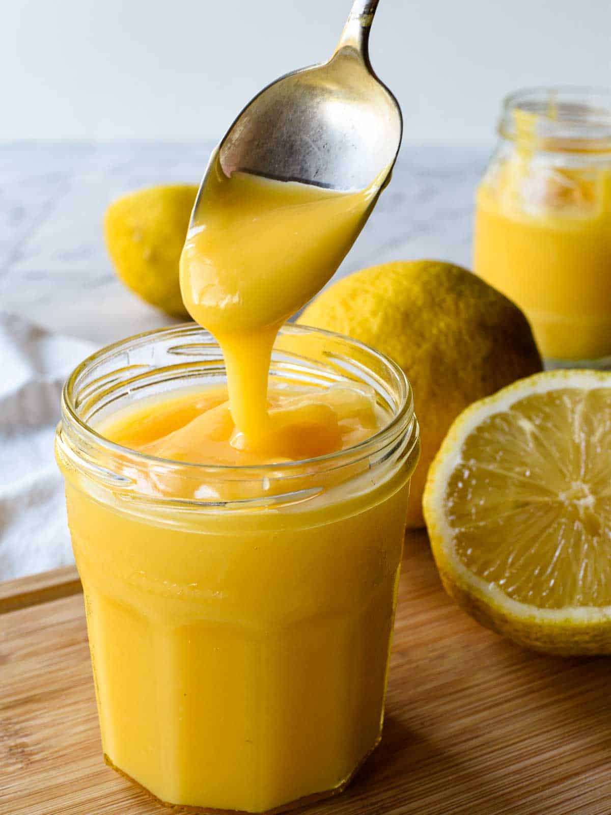Lemon curd being spooned into a in glass jar with sliced and whole lemons in the background.
