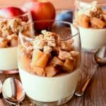 Three glasses of panna cotta topped with caramelised apples and crumble; two red apples in the background