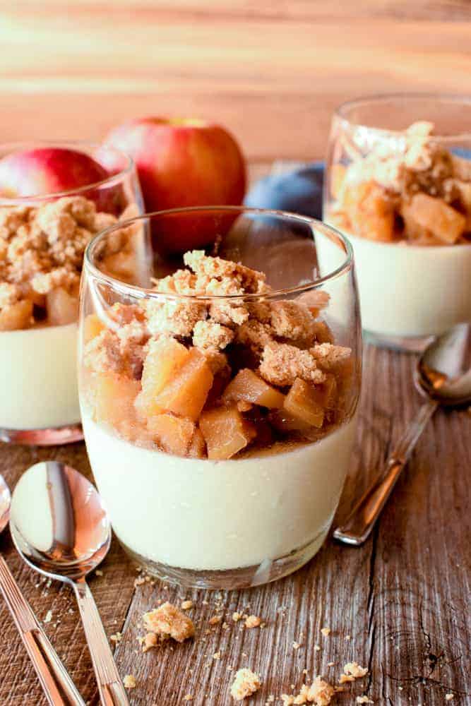 Three glasses of panna cotta topped with caramelised apples and crumble; two red apples in the background