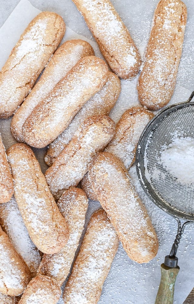 pile of oblong cookies dusted with powdered sugar and sieve on the side viewed from above.