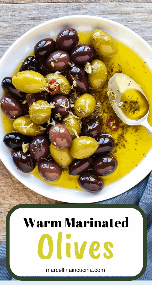 Warm marinated olives are a delicious addition to an antipasto plate. Plus they are so easy to make yourself with this simple recipe. #marinatedolives #antipasto #warmolives #spicyolives #homemadeolives white plate with assorted olives in olive oil and a spoon viewed from above