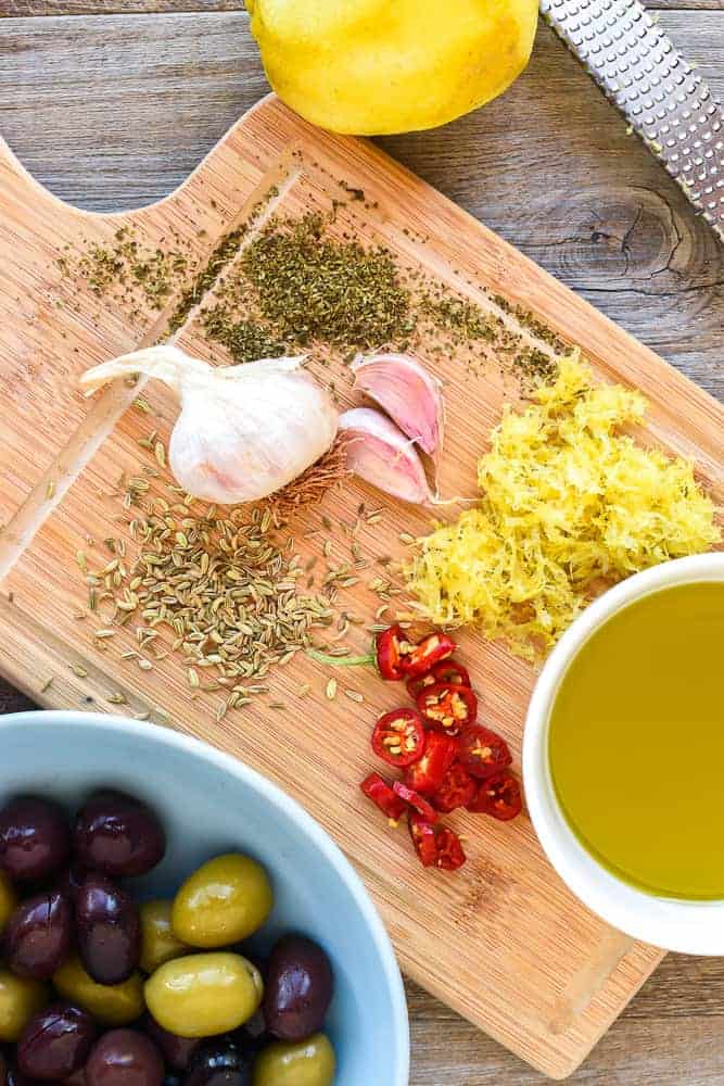 Warm marinated olives are a delicious addition to an antipasto plate. Plus they are so easy to make yourself with this simple recipe. #marinatedolives #antipasto #warmolives #spicyolives #homemadeolives chilli, dried herbs, garlic, olive oil, lemon zest, zested lemon on a wooden board with olives, zested lemon and zester viewed from above