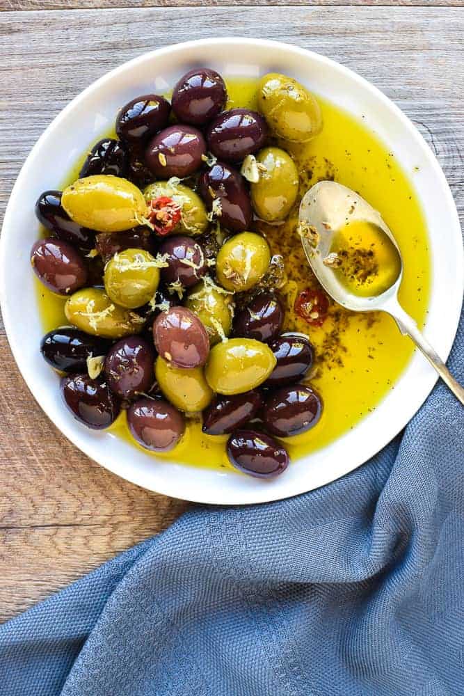 Warm marinated olives are a delicious addition to an antipasto plate. Plus they are so easy to make yourself with this simple recipe. #marinatedolives #antipasto #warmolives #spicyolives #homemadeolives white plate with assorted olives in olive oil and a spoon viewed from above