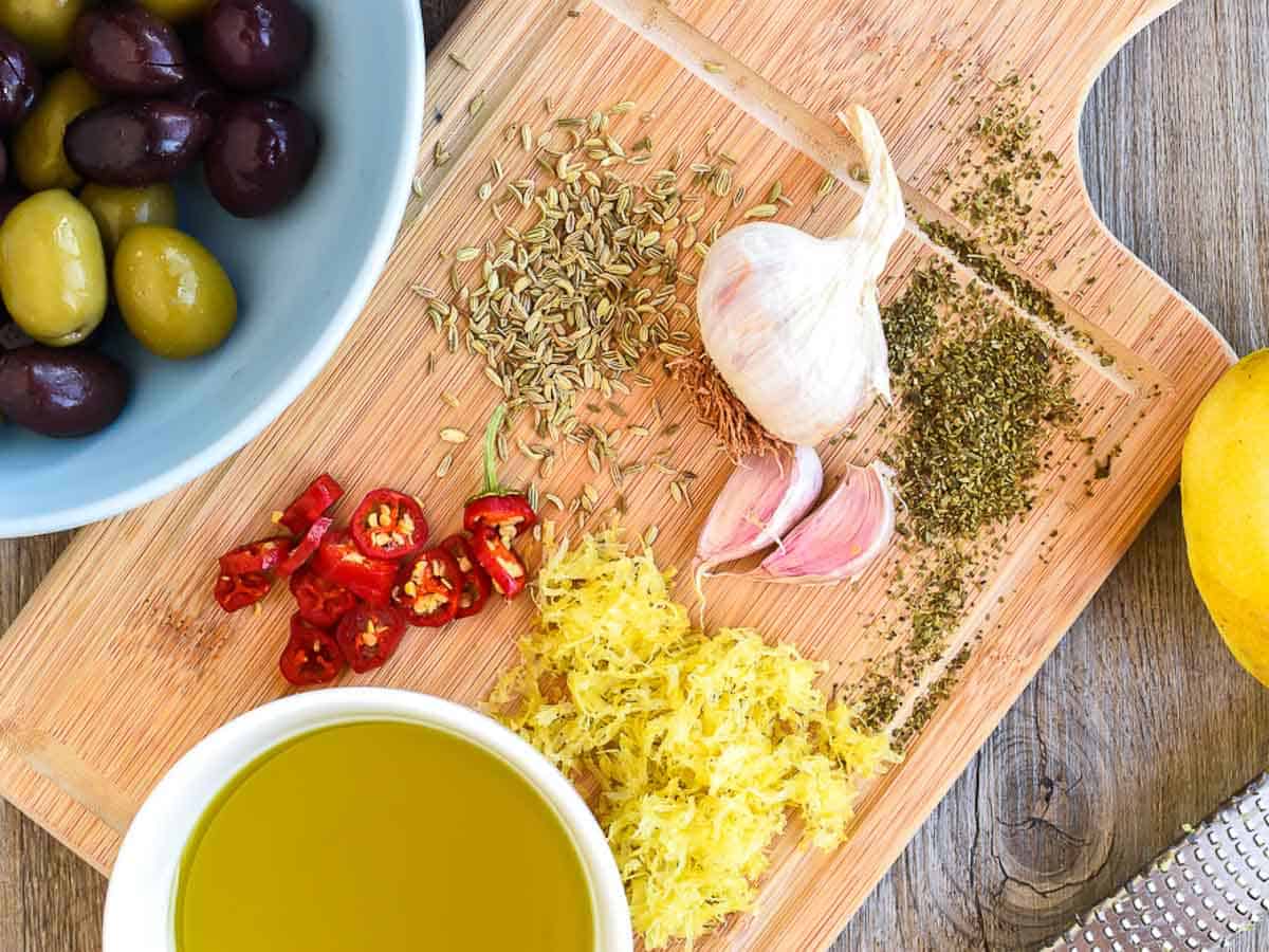 chilli, dried herbs, garlic, olive oil, lemon zest, zested lemon on a wooden board with olives, zested lemon and zester viewed from above.