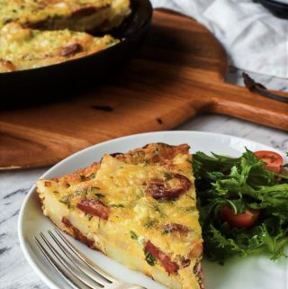 slice of potato frittata on white place with lettuce and tomato. black frypan of frittata in the background
