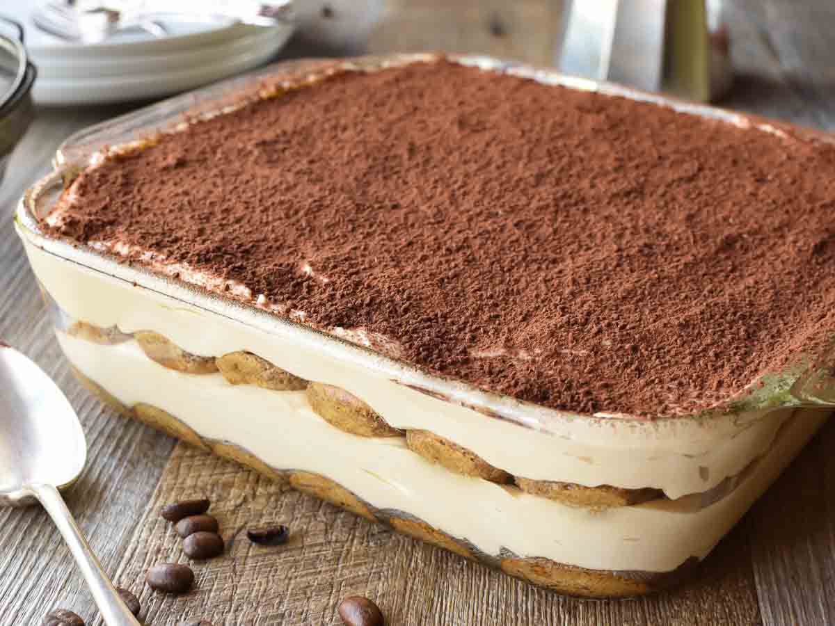 Cocoa topped, creamy dessert in square glass pan viewed from the side.