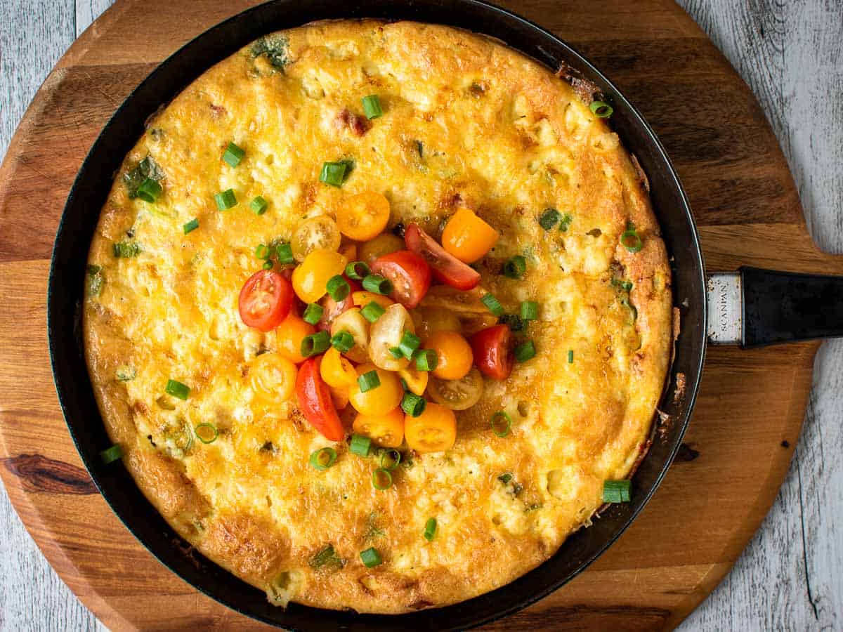 Frittata with tomato salad on top in a black frying pan viewed from above.