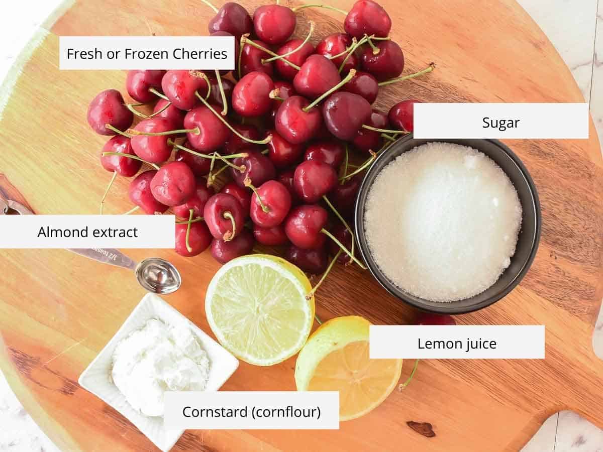 Fresh cherries, sugar, cut lemon, metal spoon and cornstarch on wooden board viewed from above.