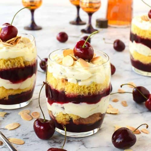 layered creamy cherry dessert in a glass with a cherry on top cherries scattered around and two other glasses of dessert in the background