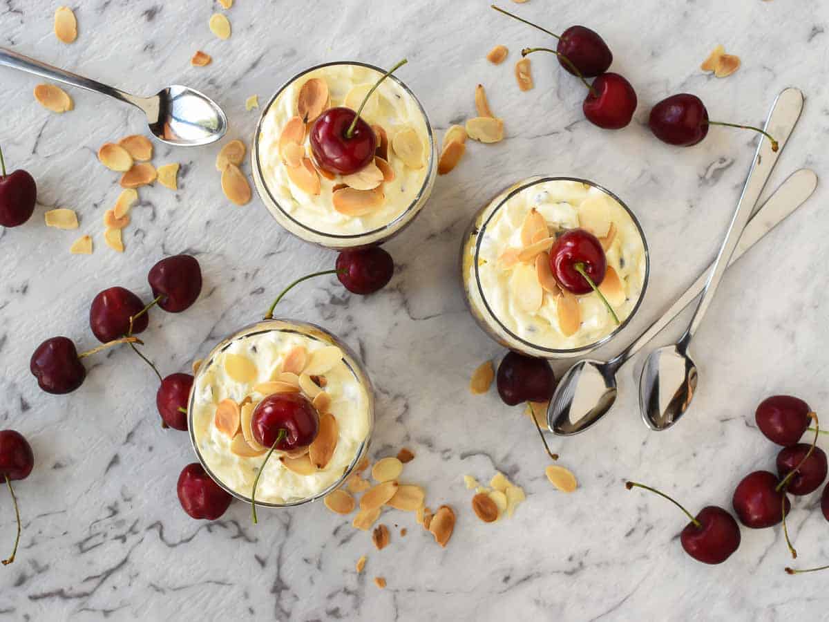 Three glasses of creamy trifle dessert with cherries scattered around viewed from above.