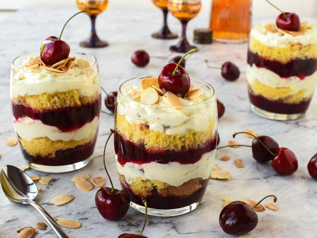 Layered creamy cherry dessert in a glass with a cherry on top cherries scattered around and two other glasses of dessert in the background.