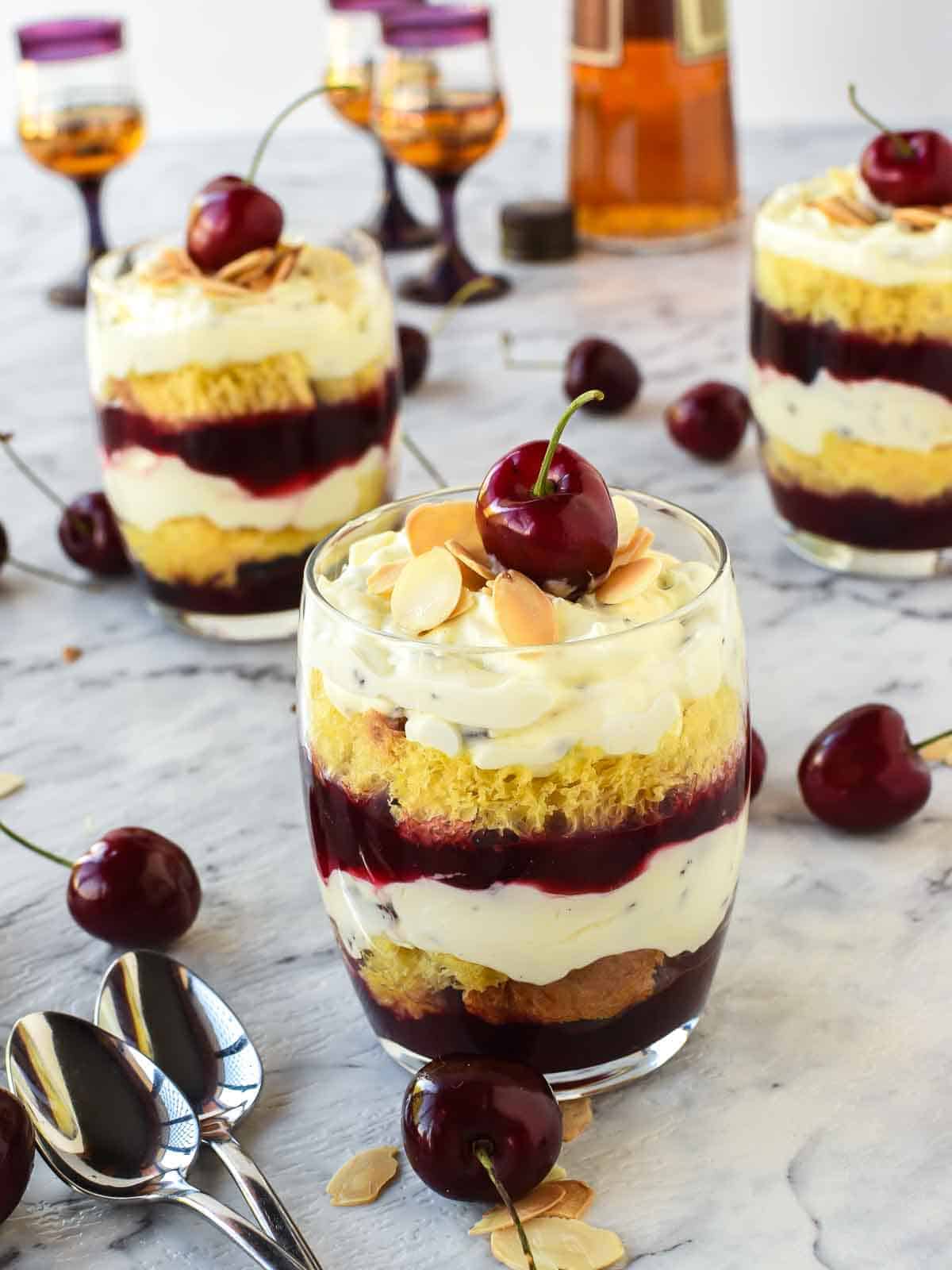 close up of mini trifles layered in a glass with a cherry on top, cherries scattered around and two other glasses of dessert in the background.