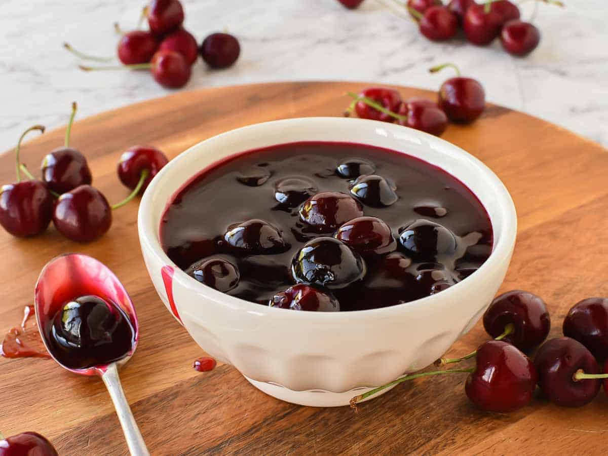 Cherry sauce in white bowl with cherries scattered around, spoonful of sauce beside bowl.