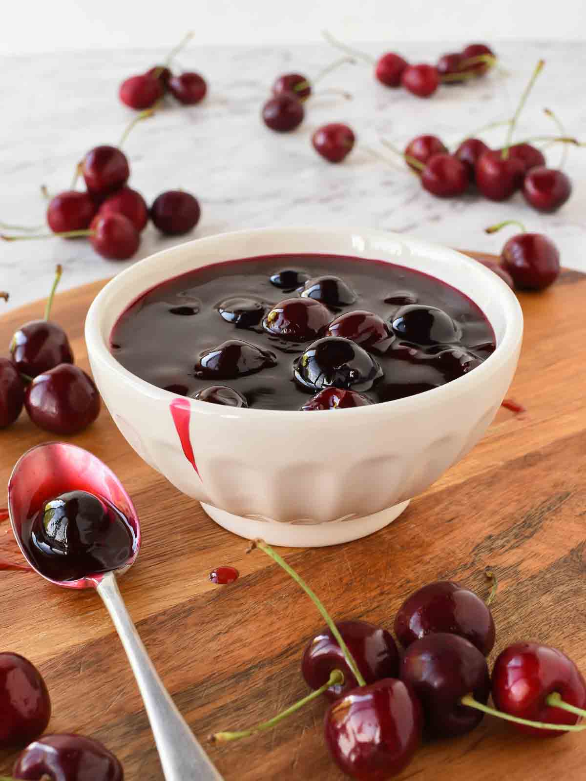 Cherry sauce in white bowl with cherries scattered around, spoonful of sauce beside bowl.