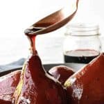4 red wine poached pears in grey shallow bowl with spoon pouring red syrup over a pear