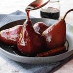 4 red wine poached pears in grey shallow bowl with spoon pouring red syrup over a pear