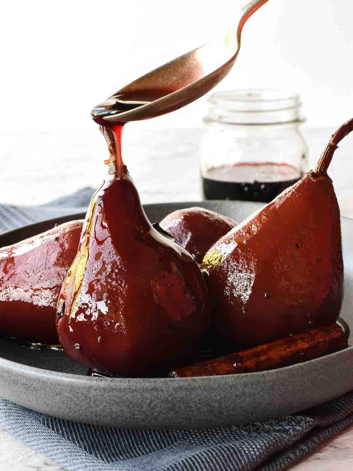 4 poached pears in red wine in a grey shallow bowl with spoon pouring red syrup over a pear