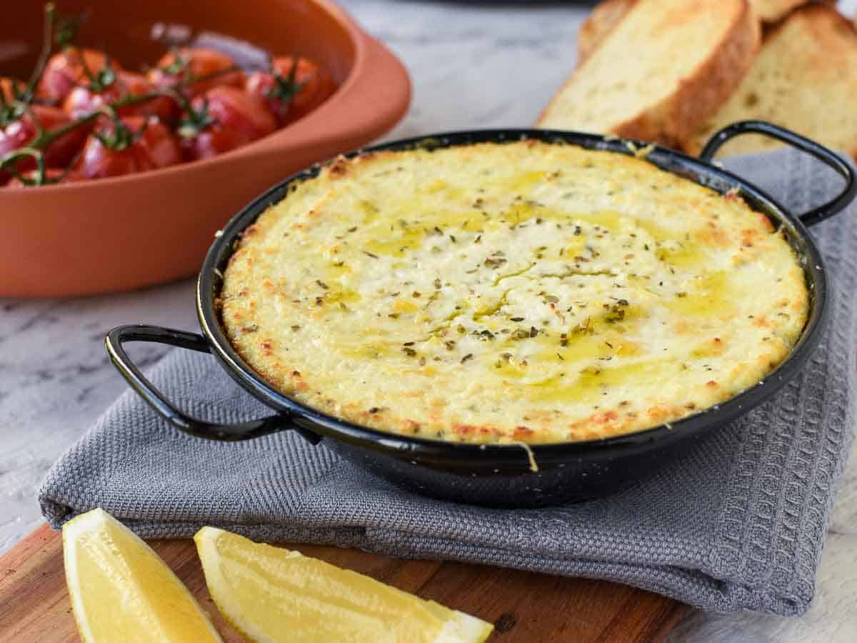 creamy baked cheese in black enamel pan on grey cloth - roasted tomatoes in the background.