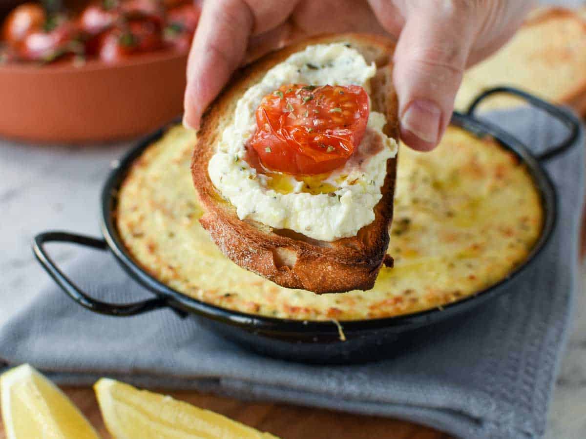 slice of toasted bread spread with white creamy dip and topped with half a slice of roasted cherry tomato.