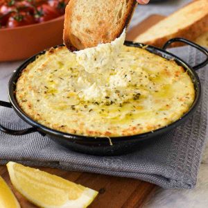 baked ricotta in black enamel pan on grey cloth with a slice of toasted bread dipping in